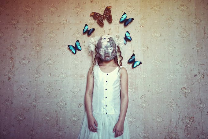 A surreal, artistic photograph of a young girl in a white dress with an old man's face overlaid where her head should be, surrounded by blue butterflies against a vintage patterned wallpaper background.