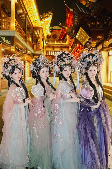 Four young women dressed in elegant pastel hanfu-style gowns with ornate floral headdresses stand together in front of a warmly lit traditional Chinese-style building, each holding a decorative round fan, capturing the ethereal aesthetic referenced in the hashtag #SAYMYNAME.