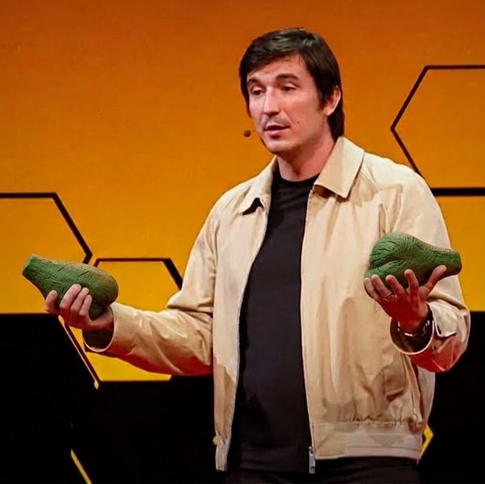A man on stage with an orange hexagonal background, wearing a beige jacket over a black shirt, holding two green, textured objects in his hands.