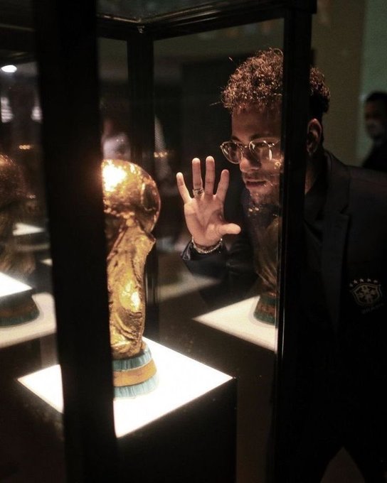 A young man with curly hair and glasses presses his hand against the glass of a museum display case, gazing intently at what appears to be the World Cup trophy, perfectly capturing the helpless, mesmerized stare of someone watching their crypto tokens vanish into the wrong address.