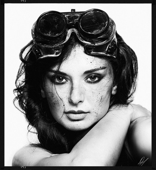 A black-and-white stylized portrait of a woman with vintage aviator goggles on her head and dark smudges on her face, with an artist's signature in the bottom-right.