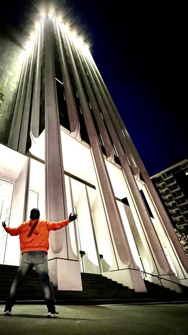 A low-angle night shot of a person in an orange hoodie standing with arms outstretched in front of a tall, illuminated Gothic-style tower.