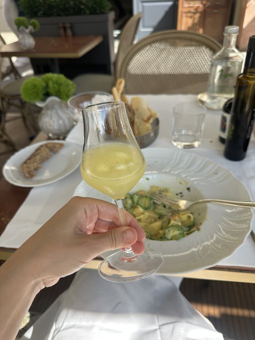A hand holds a glass of white wine over a plate of pasta at an outdoor restaurant table, with bread and water bottles also visible.