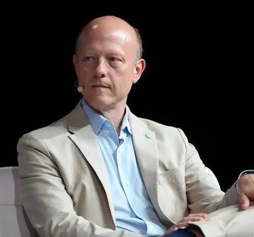 The image features a bald man wearing a light blue shirt, sitting down with his hands resting on his lap. He appears to be looking off into the distance or deep in thought. The background of the image is black, which contrasts with the white collar he's wearing and makes him stand out even more. There are no other objects or people visible in the picture, so it seems like a still photo capturing this moment.