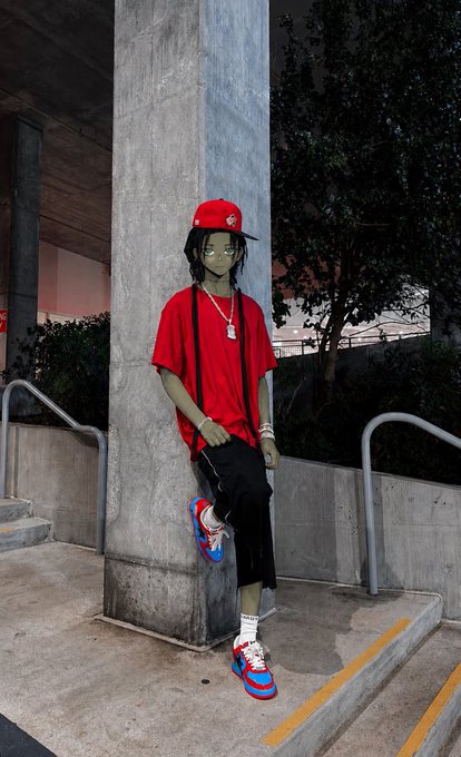 A young person wearing a red t-shirt, red cap, black pants, and red/blue sneakers leans against a concrete pillar in an urban setting.