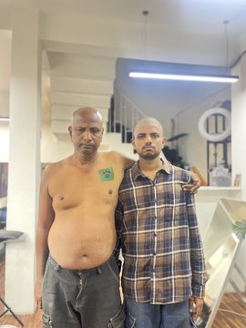 The image shows two men standing together indoors, with the older shirtless man on the left displaying a green Gamdom logo tattoo on his left chest/shoulder area, while the younger man in a plaid shirt stands beside him with his arm around the older man's shoulder. The tattoo represents the father's touching act of solidarity with his son, who has been diagnosed with cancer and is seeking support from the Gamdom team for his medical treatment expenses.