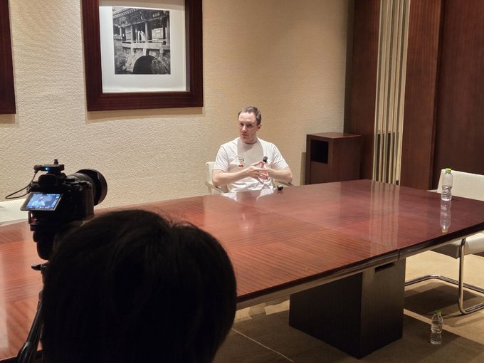 A man in a white t-shirt sits at a large conference table being filmed by a camera, appearing to give an interview or press conference.