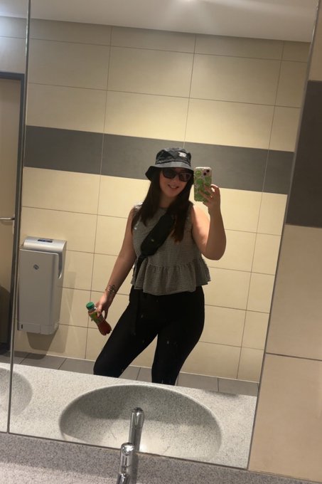 A mirror selfie of a woman in a public restroom wearing a gray top, black leggings, a black bucket hat, and sunglasses, while holding a phone with a green case and a small bottle.