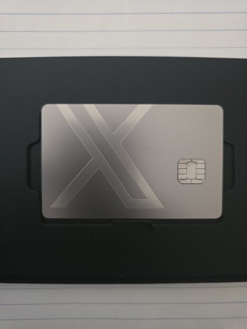 A metallic silver card holder containing an X-themed metal card with an EMV chip, resting on lined notebook paper.