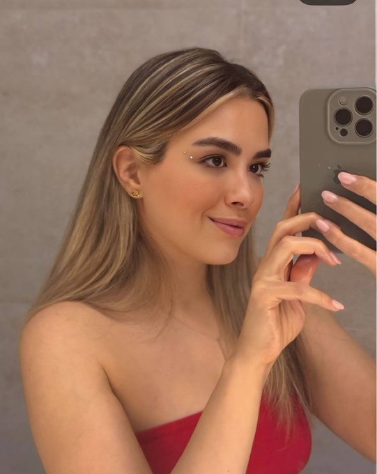 A woman with blonde-highlighted hair takes a mirror selfie with her iPhone, wearing a red top and small facial gems near her eyebrows.