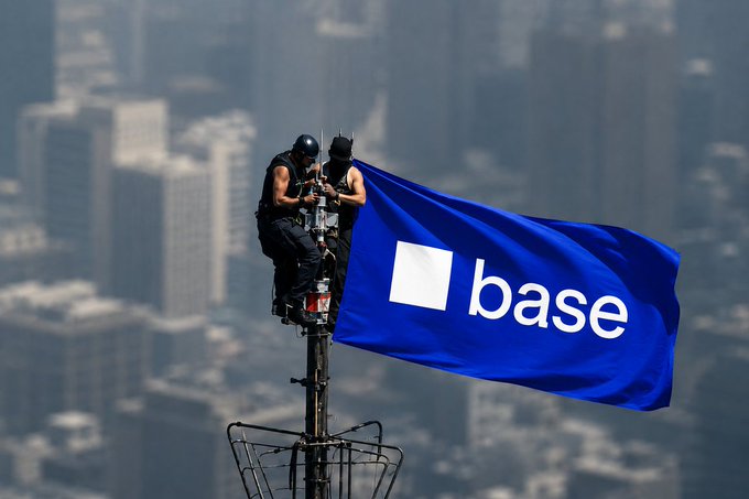 Two people on a tall antenna or tower with a city skyline behind them, holding a large blue flag with the 'base' logo (a white square next to the word 'base').