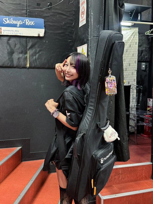 A smiling young woman with black and purple hair poses next to her large black Planets guitar case outside the Shibuya-Rex venue, suggesting she's a musician or idol performer sharing a moment of excitement about growing fan interest in "Base Tsukino."