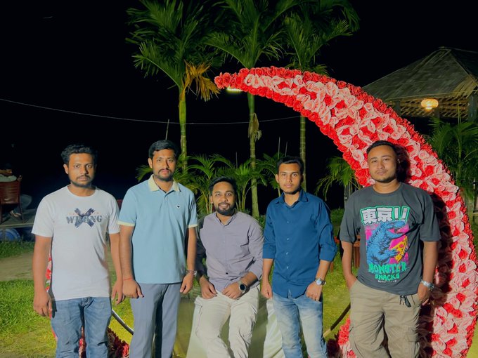 A nighttime photograph of five men posing in front of a red and white floral archway. Palm trees and a thatched-roof structure are visible in the background.