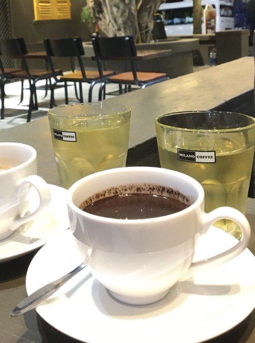 Two cups of dark coffee in white Milano Coffee cups with two glasses of iced green drinks on a cafe counter, with chairs visible in the background.