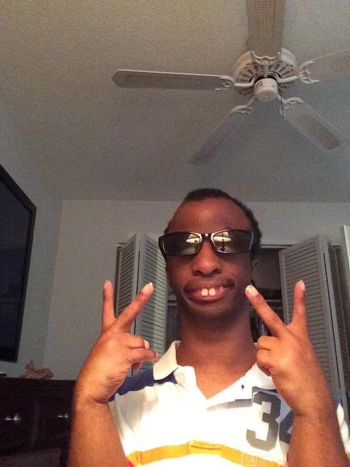 A young man wearing black sunglasses and a white jersey with the number "32" flashes a peace sign with both hands, standing in a room with a ceiling fan overhead. The peace signs visually reinforce the "Peace" message in the caption, while the casual indoor setting and friendly pose complement the upbeat "Gm" (good morning) greeting.