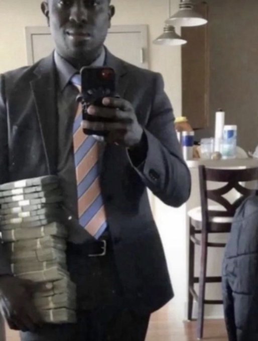 A mirror selfie of a man in a dark suit and striped tie holding a large stack of cash bands while taking a photo with his phone.