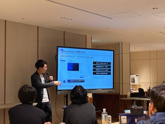 A man in a suit stands in a conference room presenting on a screen showing the SmartRender platform's actual rendering features in Japanese, while audience members watch from their seats.