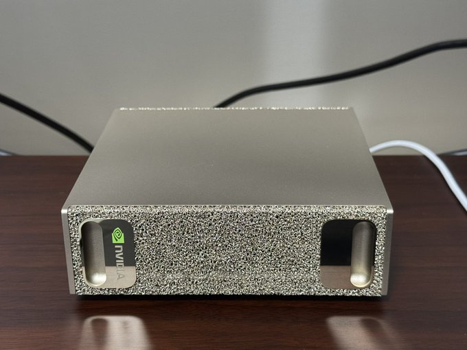 A silver, textured NVIDIA-branded hardware device, possibly a Shield or set-top box, sits on a dark wooden surface with cables connected to it.