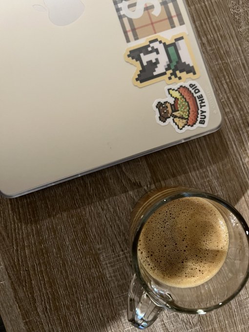 A glass mug of espresso sits on a wooden table next to a laptop adorned with pixel-art stickers, including one of a character taco.