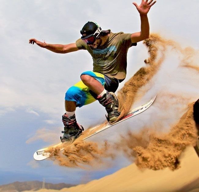 A snowboarder in mid-air performing a trick on sand dunes, kicking up a cloud of sand while wearing a green t-shirt, blue shorts, and a black cap.