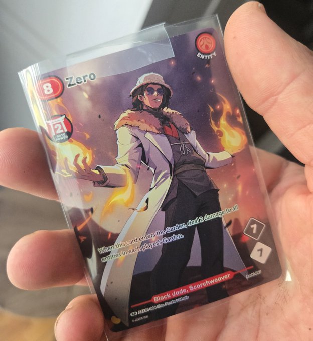 A hand holds a sealed trading card sleeve featuring 'Zero' from the Entity TCG, depicting the character 'Black Jade, Scorchweaver' surrounded by flames, with stats showing 8 power and 2 pierce.