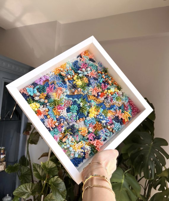 A hand holding up a white-framed shadowbox containing a colorful coral reef artwork made of beadwork/embroidery with various textures resembling sea anemones and coral, set against a backdrop of green houseplants.