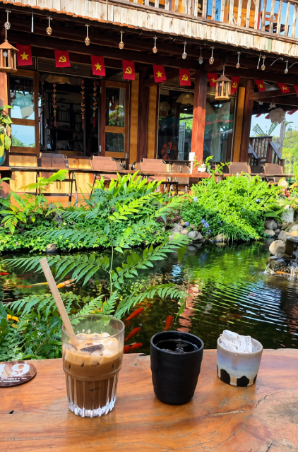 A scenic view from a wooden table holding a glass of iced coffee and small pots, looking out onto a koi pond surrounded by greenery and a traditional wooden building adorned with red flags.
