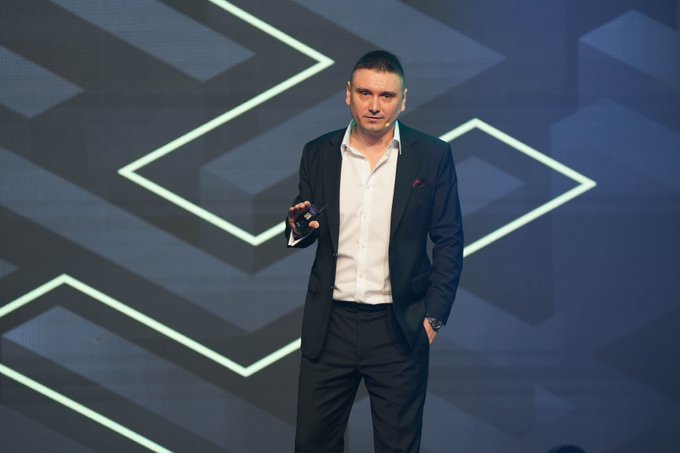 A man in a dark suit stands on stage holding a small device, appearing to deliver a presentation against a backdrop of geometric chevron patterns, likely discussing the rapidly expanding landscape of dollar-backed stablecoins.