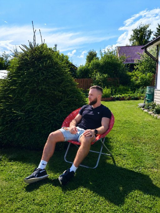 A man with a short beard, wearing a black t-shirt and light blue shorts, relaxes in a red folding lawn chair in a sunny green backyard.