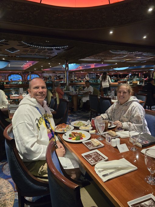 A couple is seated at a restaurant table, smiling at the camera, with plates of food and menus visible in front of them.