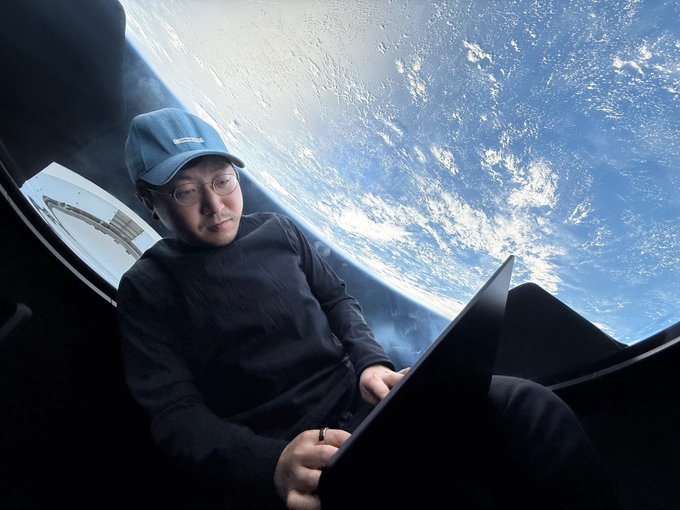 A person wearing a blue cap and glasses sitting inside a spacecraft, looking at a laptop with a view of Earth from space in the background.