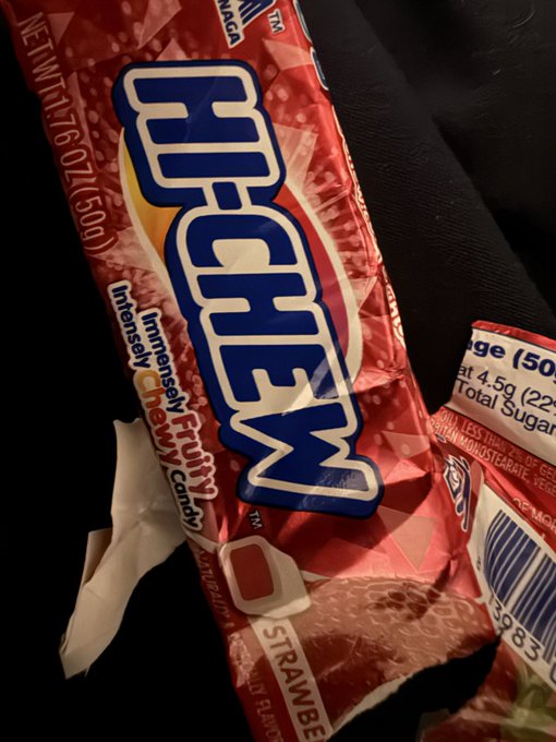 A close-up photograph of a red Hi-Chew strawberry candy wrapper with 'NEW WT 1.76 OZ (50g)' visible, alongside a partial view of a nutritional label on a dark surface.