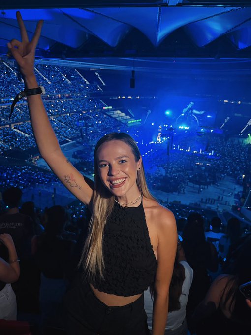 A smiling young woman with long blonde hair, wearing a black top, joyfully raises her arm in a peace sign at a large concert arena filled with blue-lit crowds and glow sticks, perfectly capturing the celebratory spirit of her birthday wish to be followed by @world_xyz.