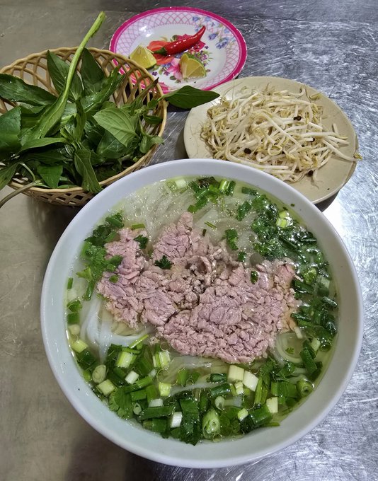 A steaming bowl of Vietnamese pho topped with sliced beef, rice noodles, and fresh green onions, served alongside a basket of herbs, bean sprouts, lime, and chili on a metal table. The vibrant presentation complements the post's warm invitation to enjoy a cozy dinner, capturing the simple pleasure of a comforting meal.