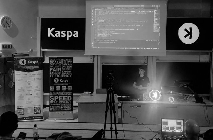A black-and-white photo of a Kaspa presentation with banners promoting decentralization, scalability, and fair launch, with a presenter standing before a projected terminal screen.
