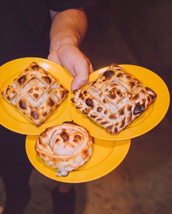 A person in a suit holding three yellow plates displaying various golden-brown baked pastries, possibly burek or similar filled dough items.