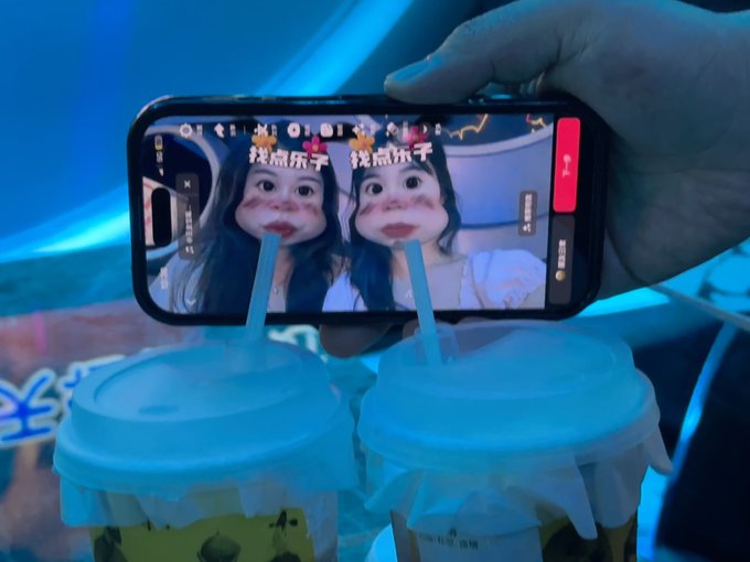 A hand holds a smartphone taking a picture of two bubble tea drinks, with the phone screen showing a camera filter featuring two cartoon faces with exaggerated lips and Chinese text '找点乐子'.