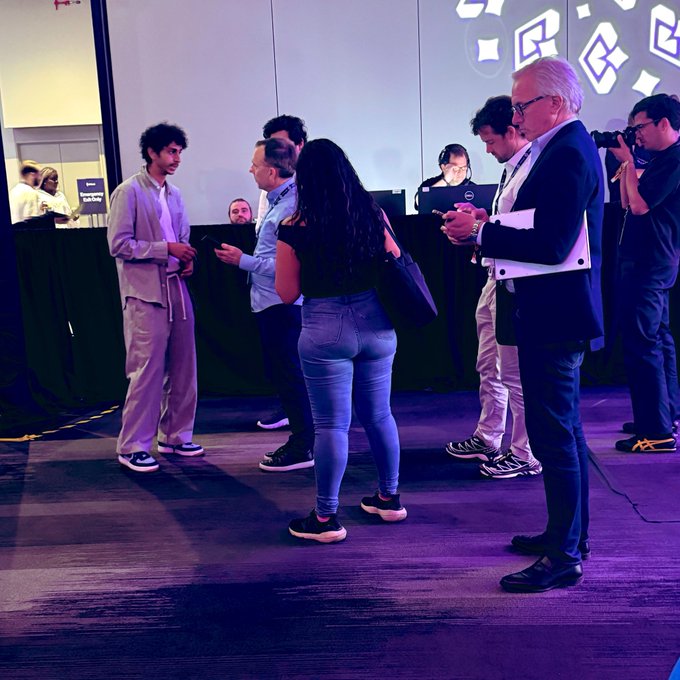 In this image, a group of people are gathered around a booth with a laptop computer. The booth appears to be set up for an event or conference related to cryptocurrency or blockchain technology. There are several individuals standing near the booth, some holding cell phones in their hands and others engaged in conversation. A man wearing a suit is also present at the booth. The people seem interested in what's being offered by the booth, possibly discussing or learning about the topic of interest.