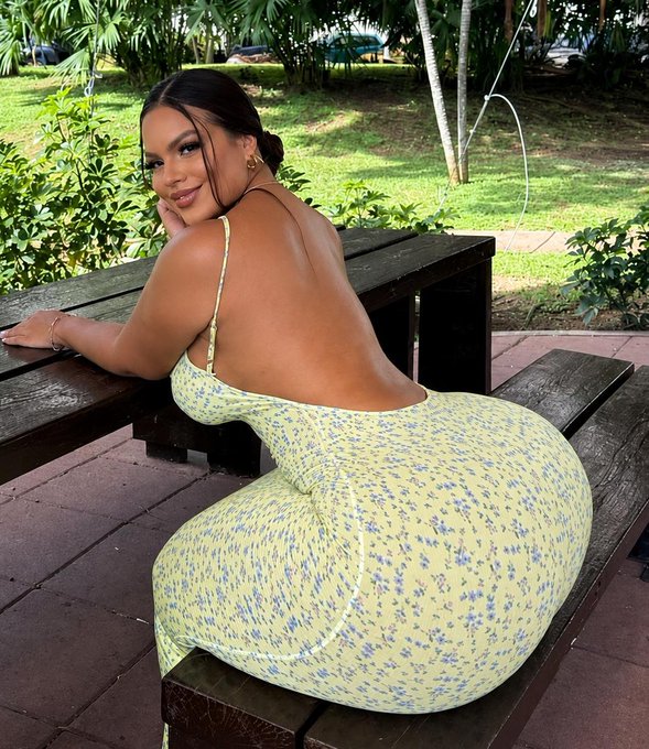 A woman with dark hair pulled back sits on a wooden picnic bench, looking over her shoulder while wearing a backless yellow dress with a blue floral print.
