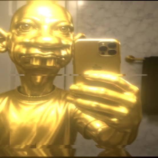 A gold-painted figure with a wide, toothy grin and large eyes takes a mirror selfie with a gold-colored smartphone.