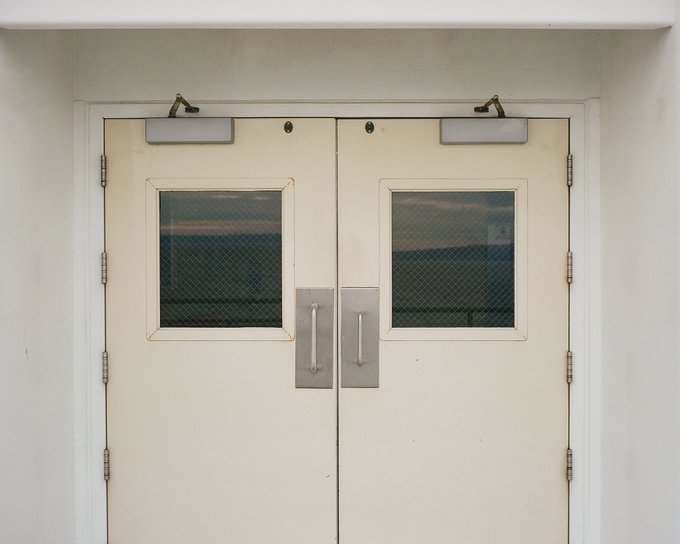 A pair of cream-colored double doors features small wire-mesh windows and silver handles in a white-walled hallway.