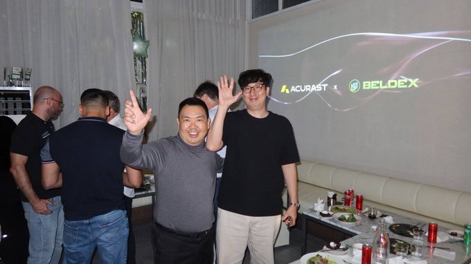 A group of people posing at an event, with two men in the center smiling and raising their hands, while a screen behind them displays logos for 'ACURAST' and 'BELDEX' in green and white text.