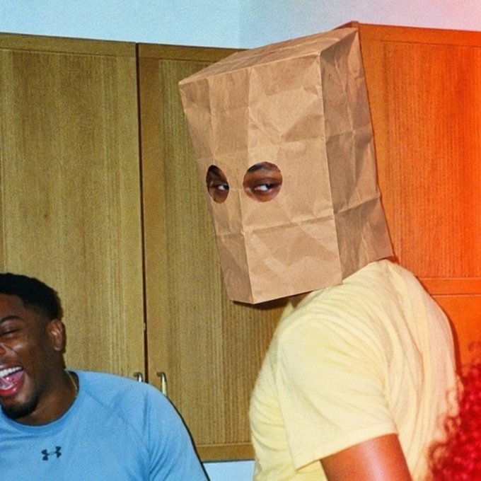 A person wearing a brown paper bag over their head with eye holes cut out, standing in a kitchen next to a man in a blue Under Armour shirt who is laughing.