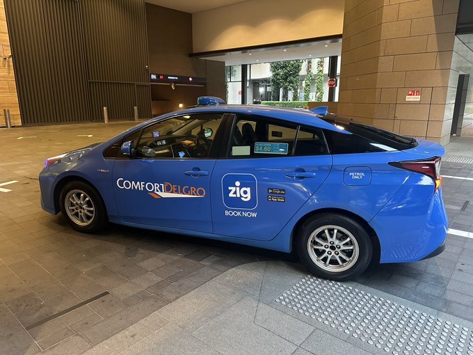 A blue ComfortDelGro Zig taxi, marked with "zig BOOK NOW" branding and a $4.60 fare displayed on its meter, is parked under a covered area at what appears to be a Singapore pickup/drop-off zone, visually reinforcing the post's reference to ComfortDelGro's ride-hailing service ($ZIG).
