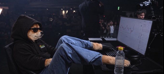 A Shiba Inu dog wearing sunglasses and a black hoodie sits casually with its feet up on a desk in front of a monitor displaying charts, appearing to be at some kind of event with people in the background.
