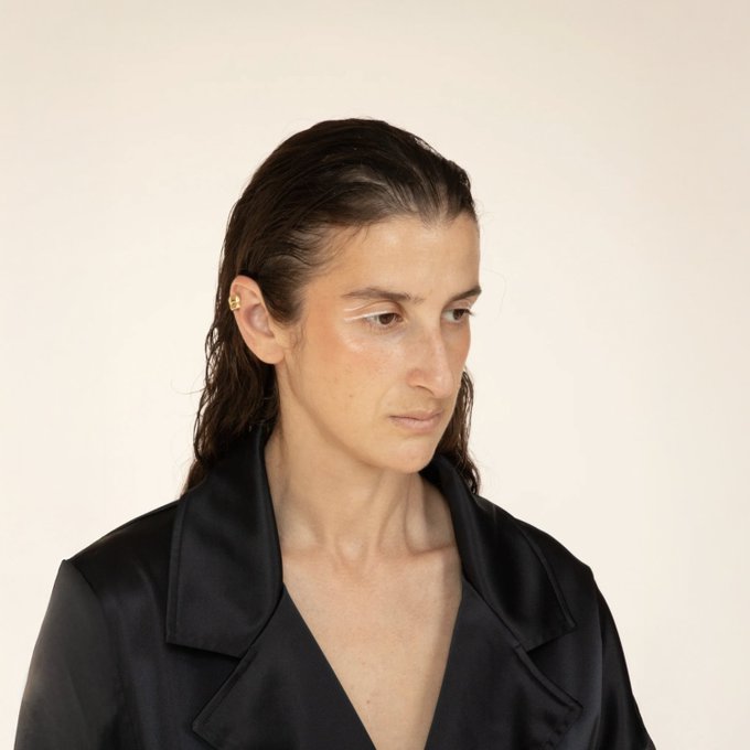 A portrait photograph of a person with wet dark hair, a small earring, and a black collared shirt against a cream-colored background.
