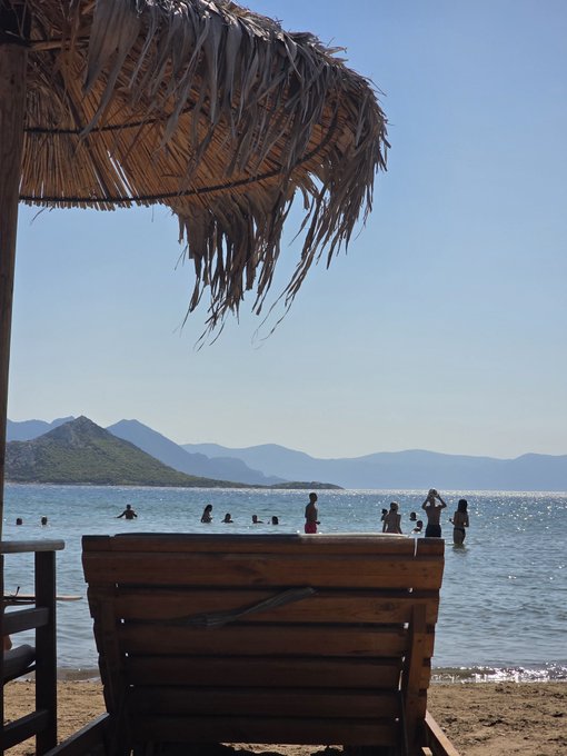 A beach scene viewed from under a thatched umbrella with a wooden chair in the foreground, showing swimmers in calm blue water with mountains in the distance.