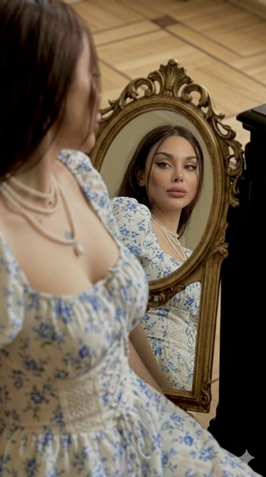 A woman in a blue and white floral dress with pearl necklaces is photographed from behind while looking into an ornate gold-framed mirror.