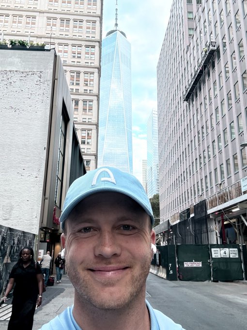 A selfie of a smiling man wearing a light blue 'C' cap, with One World Trade Center visible in the background between tall buildings in New York City.