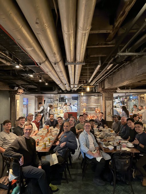 A large group of around twenty people gather for dinner at long communal tables in an industrial-style restaurant featuring exposed ceiling pipes and ductwork. The image captures the "Amp Labs x Amp dinner in Sydney" with attendees seated together amid drinks and dishes, enjoying the networking event.